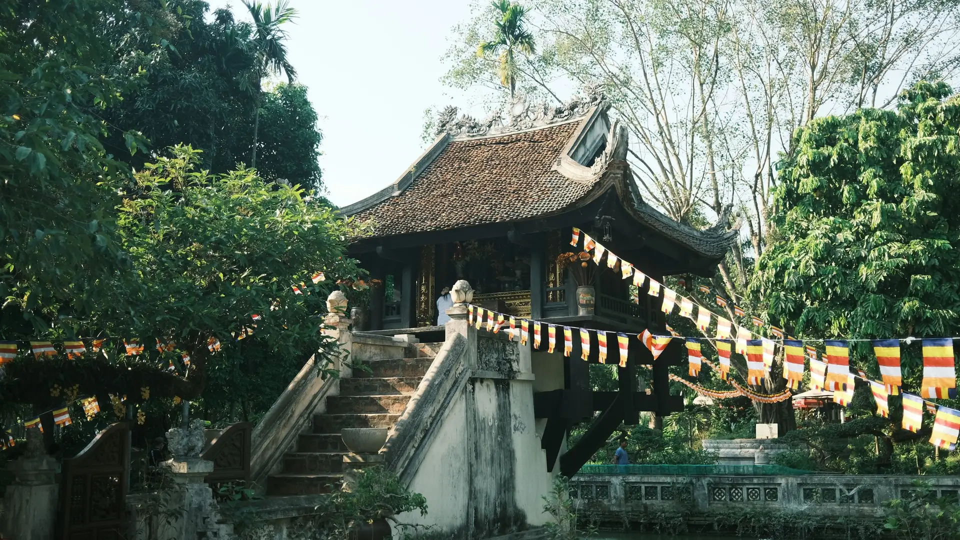 The One Pillar Pagoda in Hanoi in warm sunlight, with prayer flags and green trees