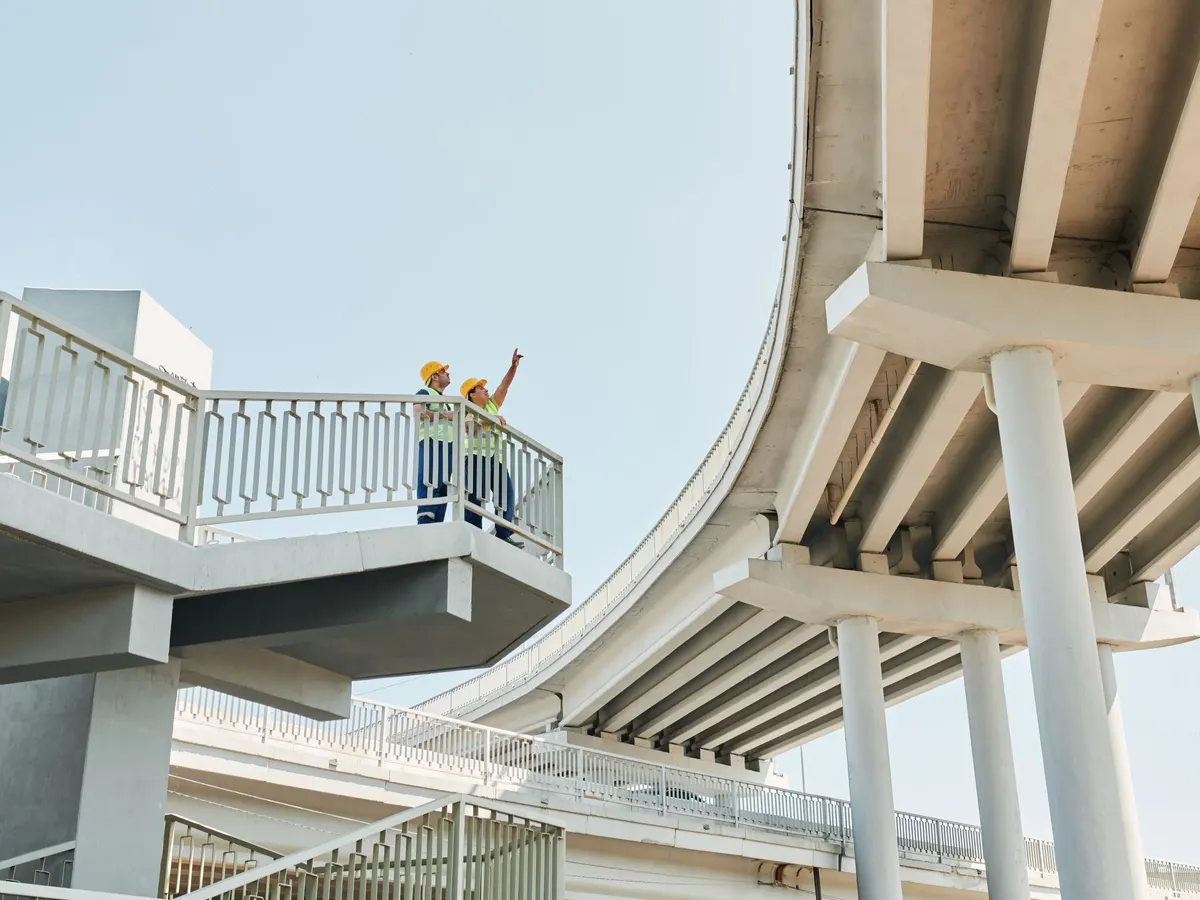 Two engineers in hard hats checking a curved concrete overpass from a walkway