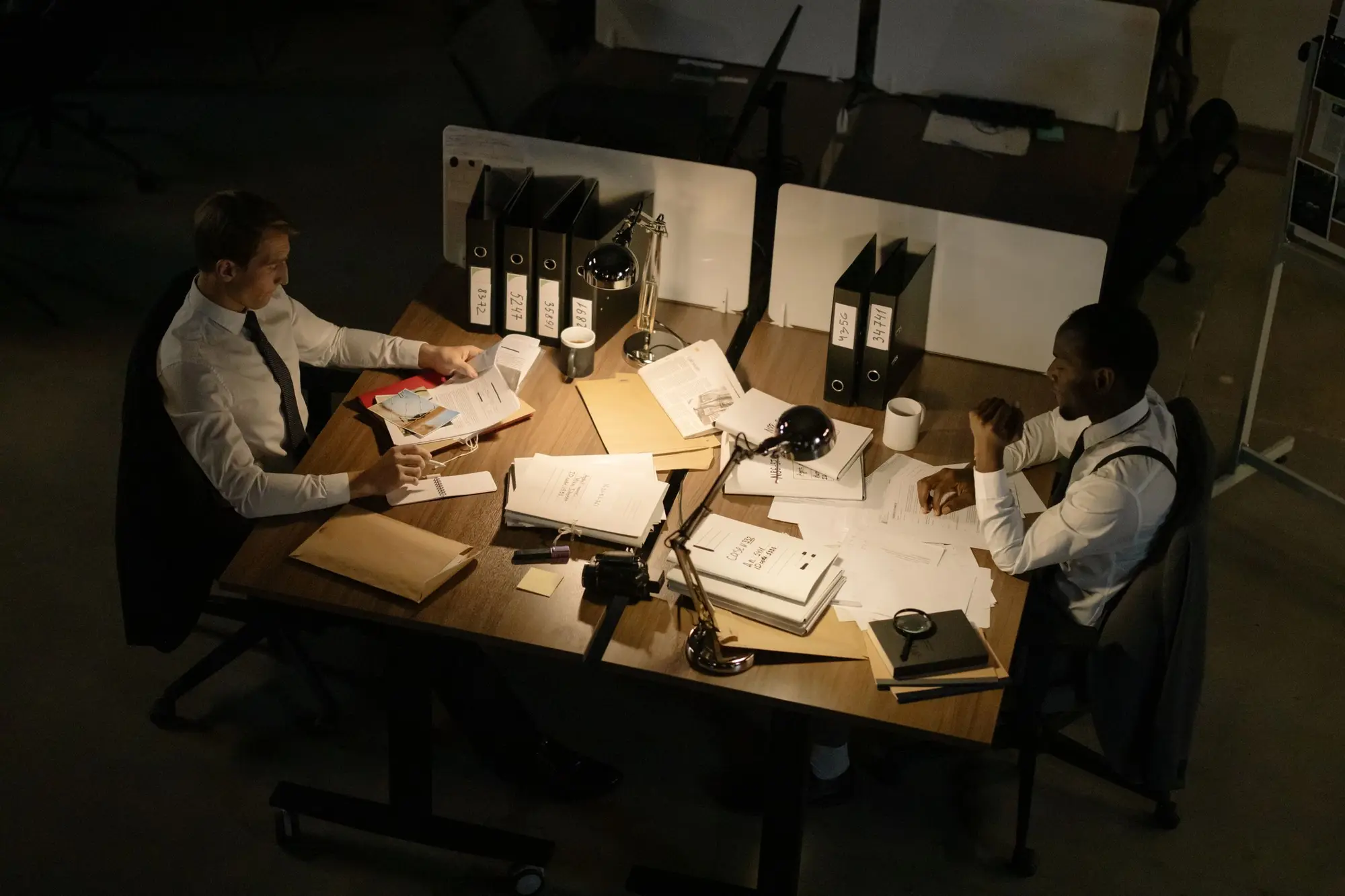 Two people at a desk covered in papers and ring binders, seen from above