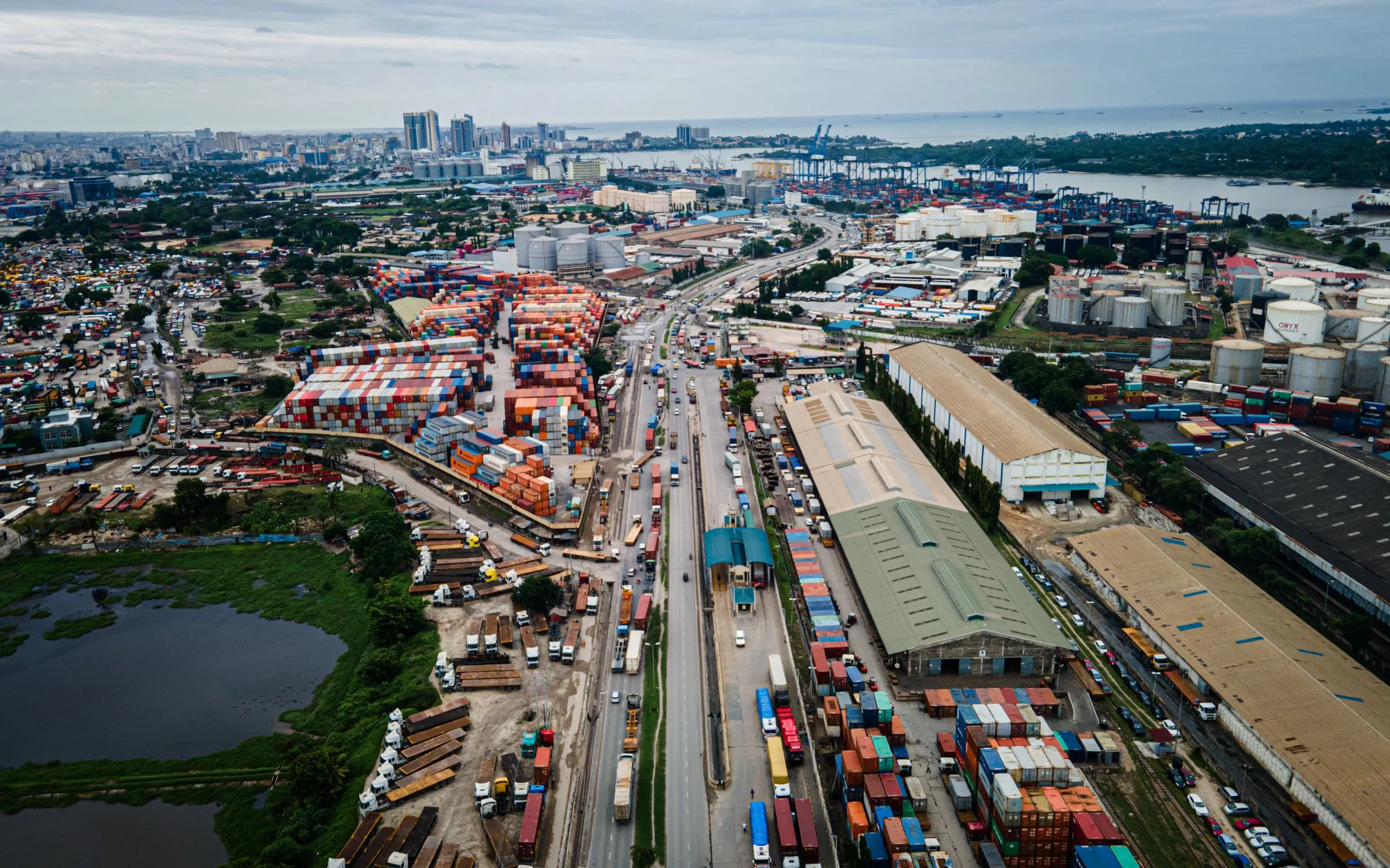 A highway lined with trucks runs from the foreground through container yards and warehouses toward a port with cranes, seen from a drone